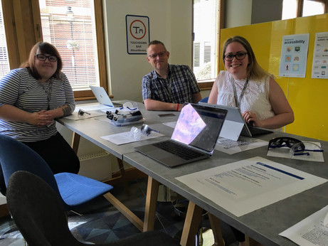 Three people seated at a high table with laptops and visual impairment simulation glasses. Behind them posters about designing for accessibility