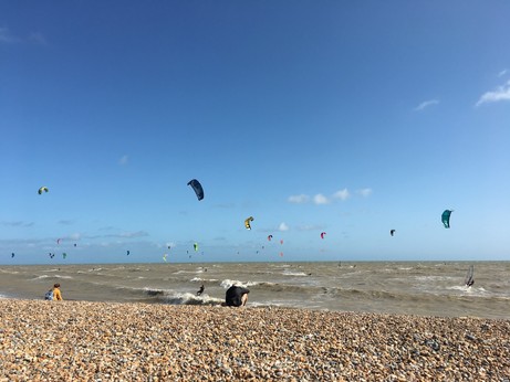 Loads of kite surfers in the sea beyond a shingle beach