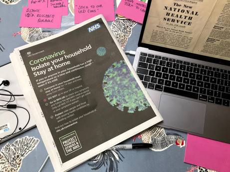 Sticky notes, laptop, headphones, newspaper with Coronavirus advert, laptop open with old NHS leaflet as screensaver