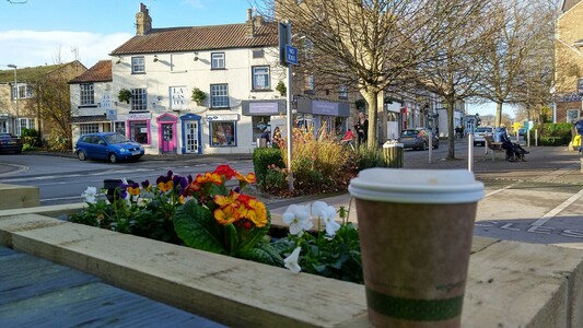 Cup of coffee resting on edge of wooden planter, in the background street and shops