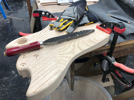 A guitar body clamped to a workshop with a Shinto rasp and some workgloves balanced upon on.