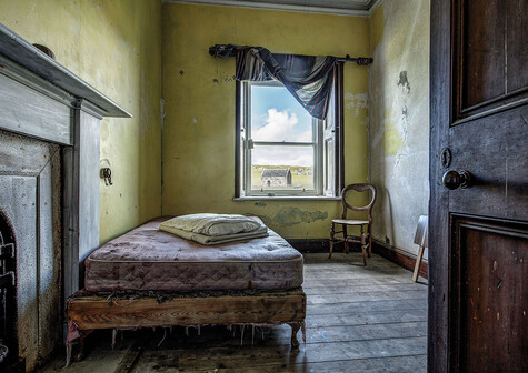 Looking into the bedroom of an uninhabited house, and out through the window to a small stone chapel in the distance