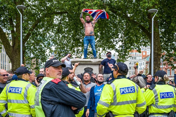 Photograph of a man waving a Union Jack flag while standing on top of the plinth where the statue of Edward Colston once stood. Around the bottom of the plinth stand a group of protestors swigging beer and chanting. Between the photographer and the protestors is a line of police in fluorescent jackets.