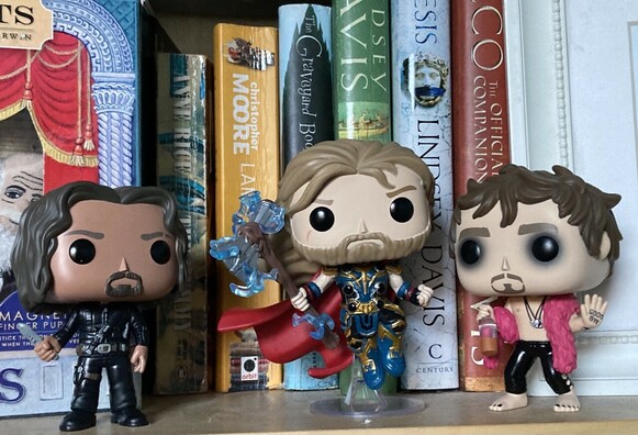Pop figures of Diego From series one of Umbrella Academy looking serious. Thor from Love and Thunder, and Klaus from Umbrella Academy season one (pink feather boa, spaced out eyes). They’re on a bookcase in front of Coraline by Neil Gaiman, a couple of Lindsay Davis titles, Lamb by Christopher Moore and The Mummy by Anne Rice.