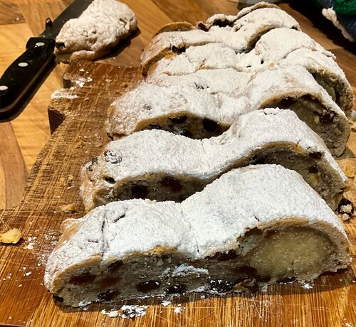 Slices of spStillen bread covered in icing. They are sat on a wooden board and you can see the line of marzipan and the fruit, There is  knife off to the left and lots of crumbs.