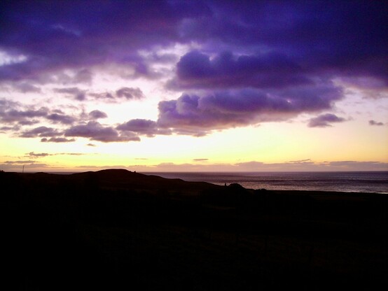 Photo of a cloudy sunset sky: Purple clouds, pale yellow and peach sky, maroon coloured sea to the left and a silhouetted hill to the foreground