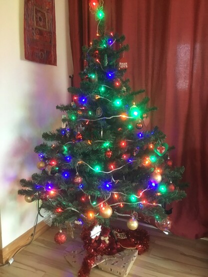 photo of a Christmas tree with lights and decorations. A red curtain behind and a cream wall at the side two gold paper wrapped presents below