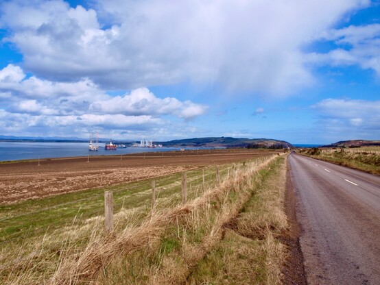 A summery photo: Blue sky with white cloud a long straight road leading towards a hill on the other side of the water (firth) which is left of the photo, dotted with oil rigs in for repair at the Nigg yard