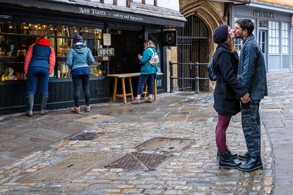 Two people, a man and woman, kiss outside a shop called Ahh Toots. It looks like it is or has been raining.