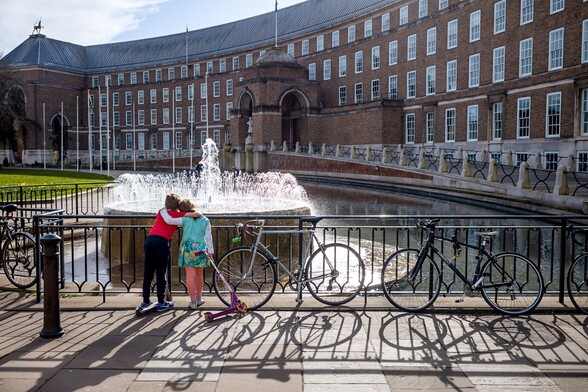 Two children look at the fountain outside of Bristol City Hall. They have arms around each others shoulders and are leaning into each other. Each has a scooter in hand.