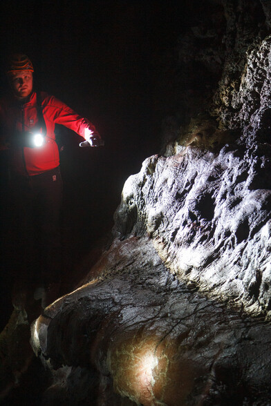 Tour guide in red jacket, hard hat, pointing at a large, rounded elephant's foot of long-solidified lava - scene illuminated only by a few flashlights, deep in an Icelandic lava tube.