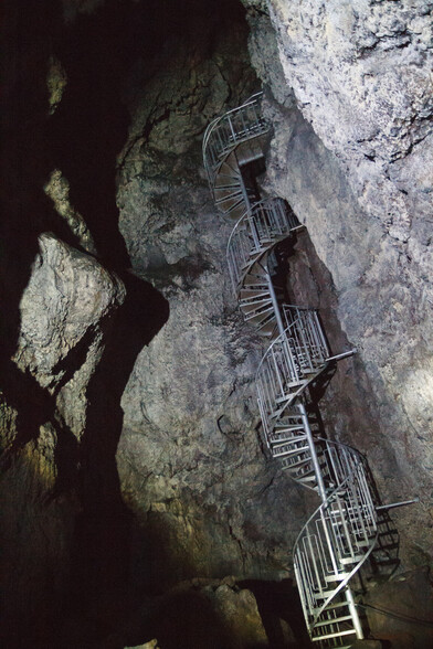 Spiral metal staircase descending deeper still underground, in a huge, lumpy volcanic chamber - seen from beneath and one side, illuminated only by bright flashlights.