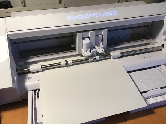 A view of the mechanism of a computer-controlled cutter. The machine is part of the way through a cut on some plastic sheet material. The machine is a light grey, and has a backlit name branding it. 