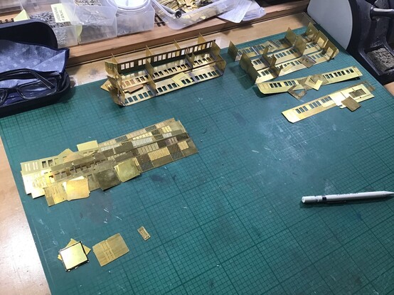 Various etched brass components to make up scale model railway coach interiors. The parts are arrayed, sorted into respective piles, on a dirty green cutting mat. There is also a propelling pencil and glasses case in the frame.