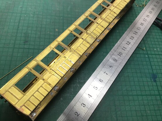 Once more, on the scruffy green cutting mat, this time with one of the brass photo etched coach bodies the handrails are for. Four mostly successful handrails are temporarily fitted in the holes in the body. There is a steel rule marked in centimetres for scale.