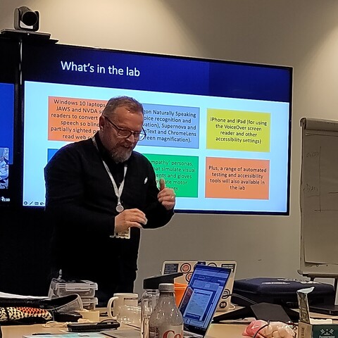 A man with glasses and a beard in front of a screen. On the screen is a PowerPoint slide titled "What's in the lab"