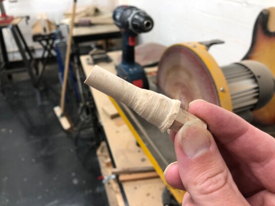 I’m holding in my hand a bit of wood that is cylindrical at the end, tapering down from a winder middle and has a narrow bit at the the other end, which I’m holding. Behind it you can see a power drill and a disk sander.