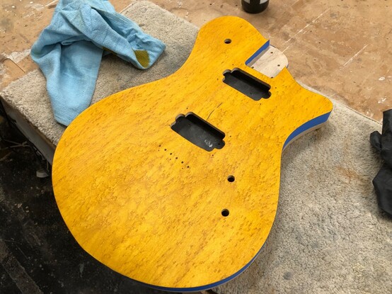 A guitar body sits on the workbench. The top of the guitar body is a figured bit of birds-eye maple and has been stained a deep (almost orange) yellow. You can see tape around the edge to stop the stain getting to the sides (hopefully).