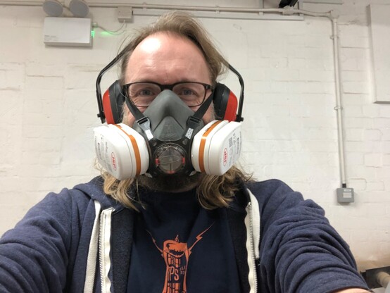 A selfie of me wearing a respirator and ear defenders and glasses in front of a painted brick wall