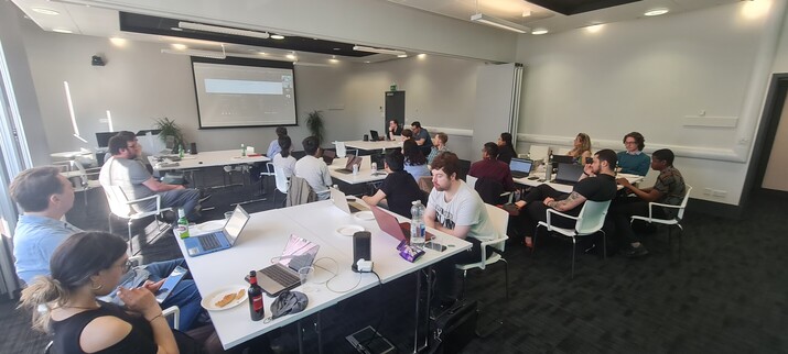 23 developers and data practitioners Sat at tables with laptops listening and watching a Zoom presentation. 