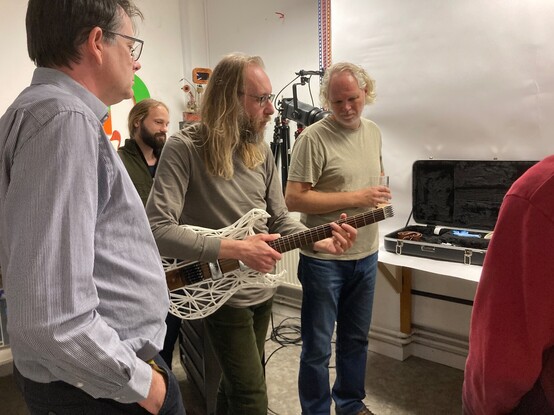 A photo of me holding my Älgen wood and 3D-printed guitar in a cluster of onlookers. I look like I've escaped from a 70s edition of the open university programs.