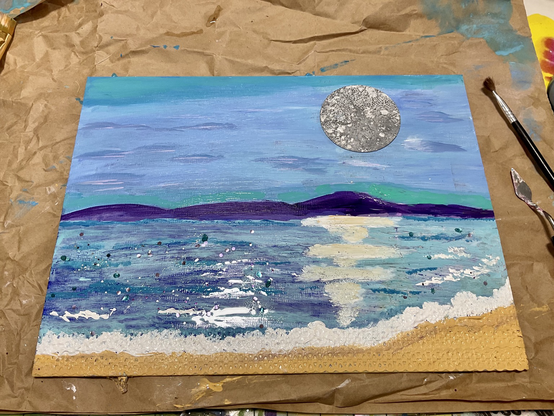 Photo of a painting of a mixed media seascape with a huge silver moon reflected in water. Purple hills and a slither of sand and surf. Glitter.