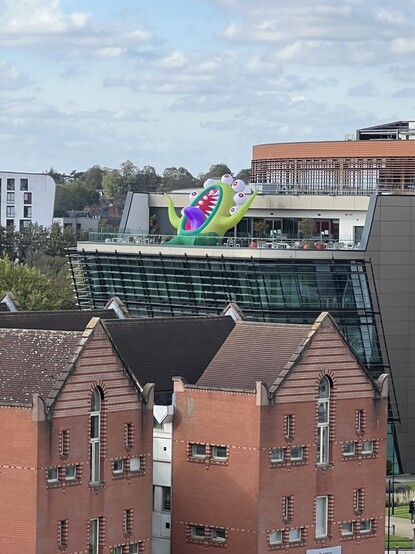 The Vijay Patel building at De Montfort University with a large inflatable monster with tentacles and many eyes as part of the Leicester city Halloween trail. It is leaning backwards making it look up at the sky, a huge mouth in the centre open with a giant purple tongue sticking out. It looks like it’s howling and waggling its arms in frustration. 