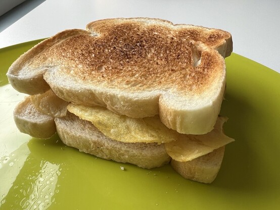 A toasted crisp sandwich resting on a green plastic plate. The crunchy edges of some cheese-and-onion crisps are poking out from between two slices of toasted white bread. Mmmmm. 