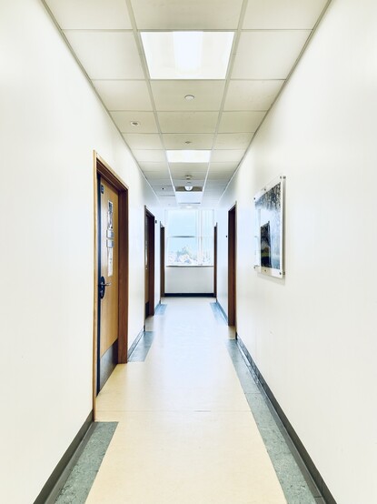 Photograph of a. Empty hospital corridor. Over exposed to increase the feeling of a bright white empty space. At the end of the corridor is a window looking out towards houses and a grey blue sky. 