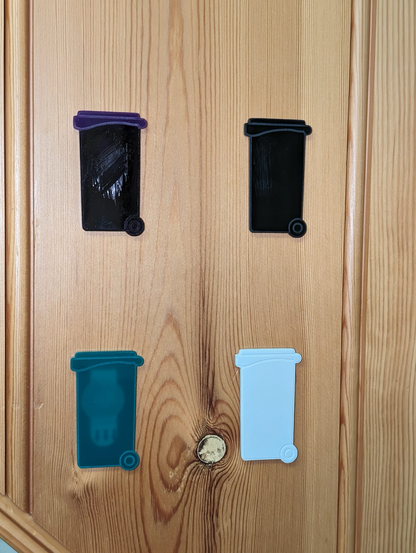 Four lasercut representations of wheelie bins, of various colours (black, grey, green, black with a purple lid) stuck to a wooden background.
The green one is semi-transparent, showing an NFC sticker, and some double-sided adhesive tape through the bin.