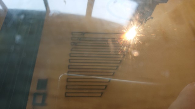 View through the inspection window of a laser-cutter at a cut in progress.

There's an array of long thin rectangles visible on the brown MDF and in the top right corner a bright flame where the cutting is happening 