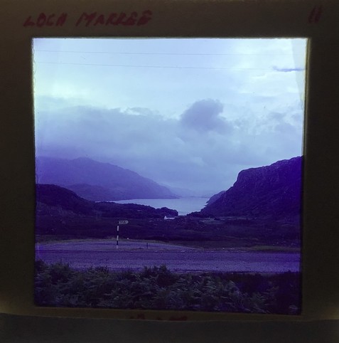 A slide titled "Loch Marree" taken in the late 1960s. A loch can be seen flanked by steep hills with clouds in the sky. In the foreground is a road and an old fingerpost with a black and white striped post. There isn't enough resolution to read it.