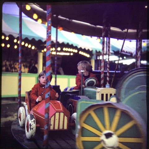 Two young children on a colourful merry go round. A young girl is in a red racing car. A younger boy is on a tractor. The background is an out of focus fairground.