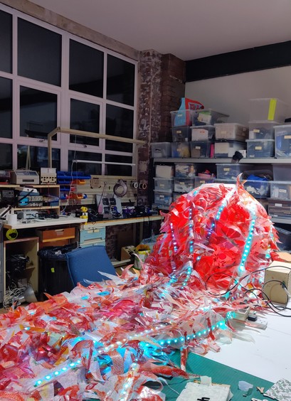 A large mediaeval-style comet with red and green addressable LEDs, with a red coma and red and green tails covered with repurposed soft plastic, lying on a white table in a maker space