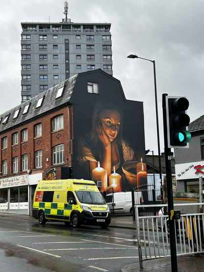 A mural on the end of a row of buildings: a girl stares at four lit candles, her chin resting on her hand in contemplation. There’s an ambulance parked on the road in front, and a grey tower block looms in the background 