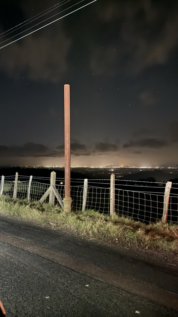 The Cinderford rusty pole landmark at night with lights along the horizon. 