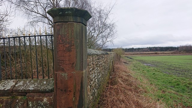 The corner of a stone wall enclosing the Earl of Derby's grounds.  

Along one edge is a low stone wall topped with iron railings; the corner is marked with a stone pillar and the remaining wall is a higher stone wall running away from the camera towards the vanishing point.