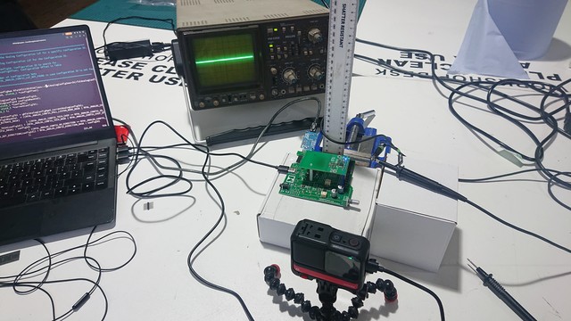 A white workbench with a laptop, oscilloscope, camera and a green circuit board on it.

The camera is pointing at the circuit board and the oscilloscope screen and a ruler which is clamped to allow measurements of height above the circuit board 