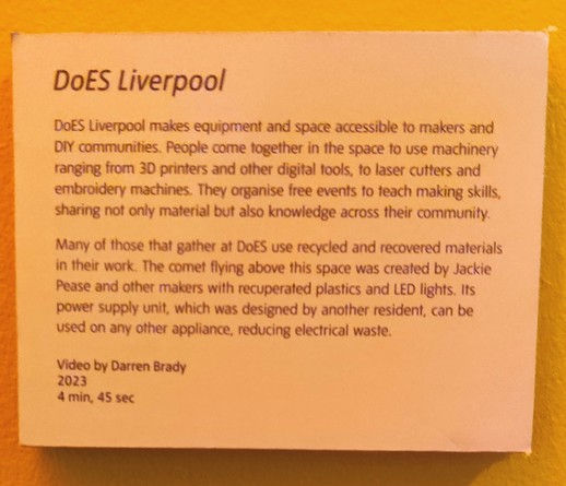 Black writing on a white background (but lit with a dull amber light) reads:
DoES Liverpool
DoES Liverpool make equipment and space available to makers and DIY communities. People come together in the space to use machinery ranging from 3D printers and other digital tools, to laser cutters and embroidery machines. They organise live events to teach making skills, sharing not only material but also knowledge with the community.

Many of those that gather at DoES use recycled and recovered materials in their work. The comet flying above this space was created by Jackie Pease and other makers with recuperated plastics and LED lights. Its power supply unit, which was designed by another resident, can be used in any other appliance, reducing electrical waste.