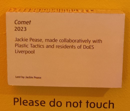 Black writing on a white background (but lit by a dull amber light) reads:
Comet 
2023
Jackie Pease, made collaboratively with Plastic Tactics and residents of DoES Liverpool

Underneath it says "Please do not touch"