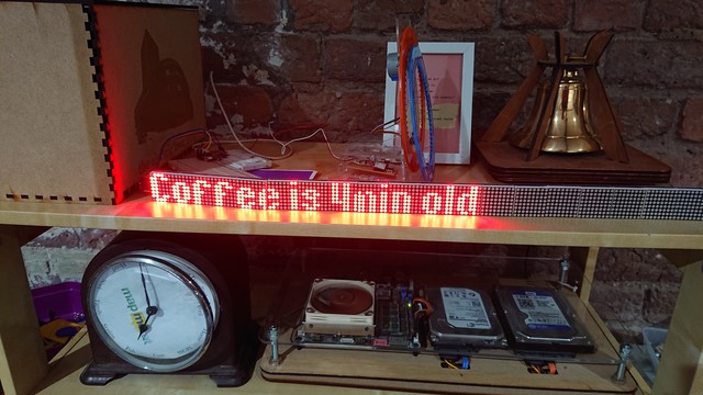 A single line dot-matrix text display sits on a shelf next to a brass bell and above a location clock and a computer.

The text on the display reads "Coffee is 4min old" 