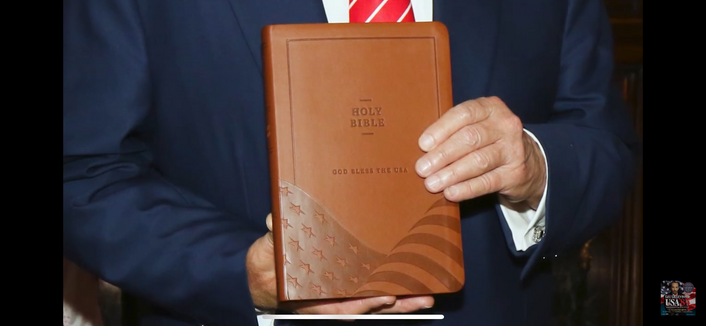 A brown Bible being helped by Trump with the words Holy Bible and God Bless the USA and an American flag on the bottom