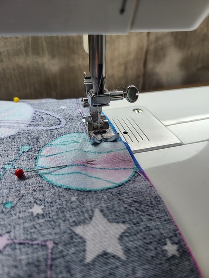 The presser foot of a white sewing machine, midway through sewing a seam. The machine is threaded with grey thread. The visible fabric is showing the "wrong" side of a space themed, planet-encrusted fabric. Two pins are visible holding fabric together, one red and one yellow.