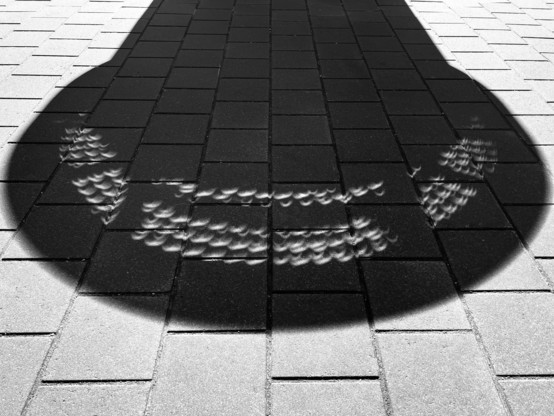 The shadow of a public artwork, its many small holes making half-moon shapes on the ground, echoes of the eclipse happening overhead 