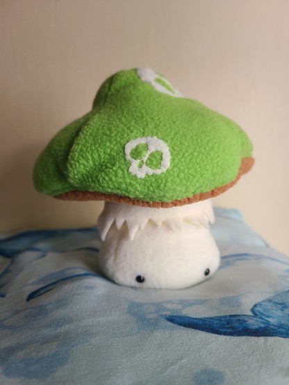 A little soft toy mushroom with eyes, sitting on a pillow with a white and blue, whale-themed pillow.

The hat is green and has skulls for spots.