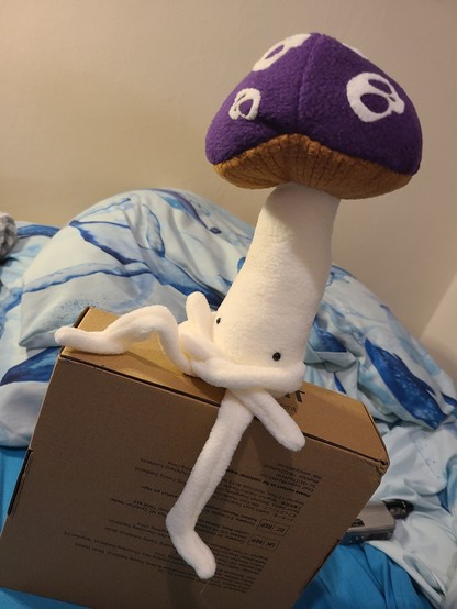 A little soft toy mushroom with eyes, sitting on a pillow with a white and blue, whale-themed pillow.

The hat is purple, with skulls for spots. One leg is up to the left, foot down and knee up. The arms are crossed.