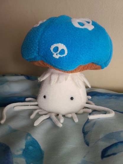 A little soft toy mushroom with eyes, sitting on a pillow with a white and blue, whale-themed pillow.

The hat is bright blue. Two long, thin arms are either side, and the mushroom has lots of little thin roots of various lengths.