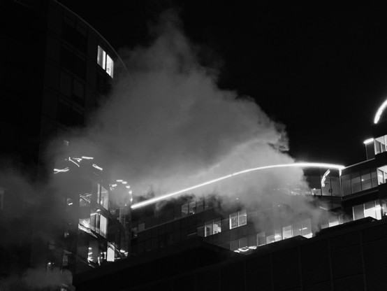 Black and white city scene, looking up - steam rising from a rooftop, neon lights and windows glowing through 