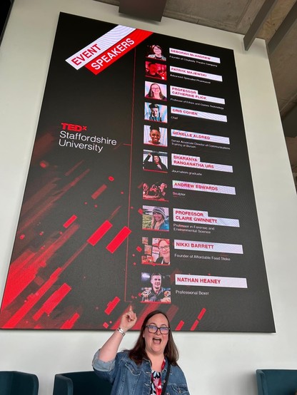 A picture of me pointing up at a sign with all the TEDxStaffordshireUniversity speakers on it (including me!)