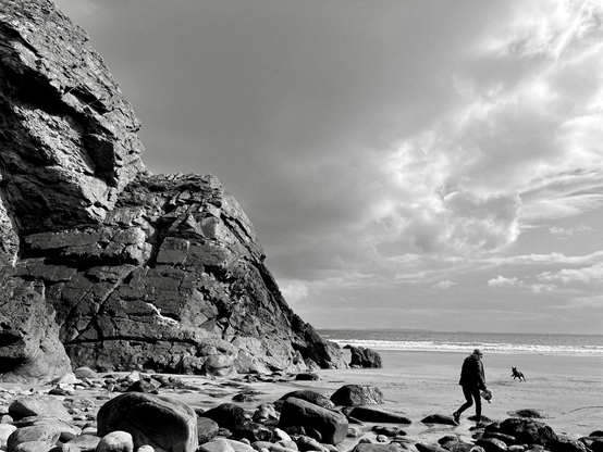 Black and white photo of Caerfai beach in Wales - steep rocks on one side. A human and a dog playing on the sand 
